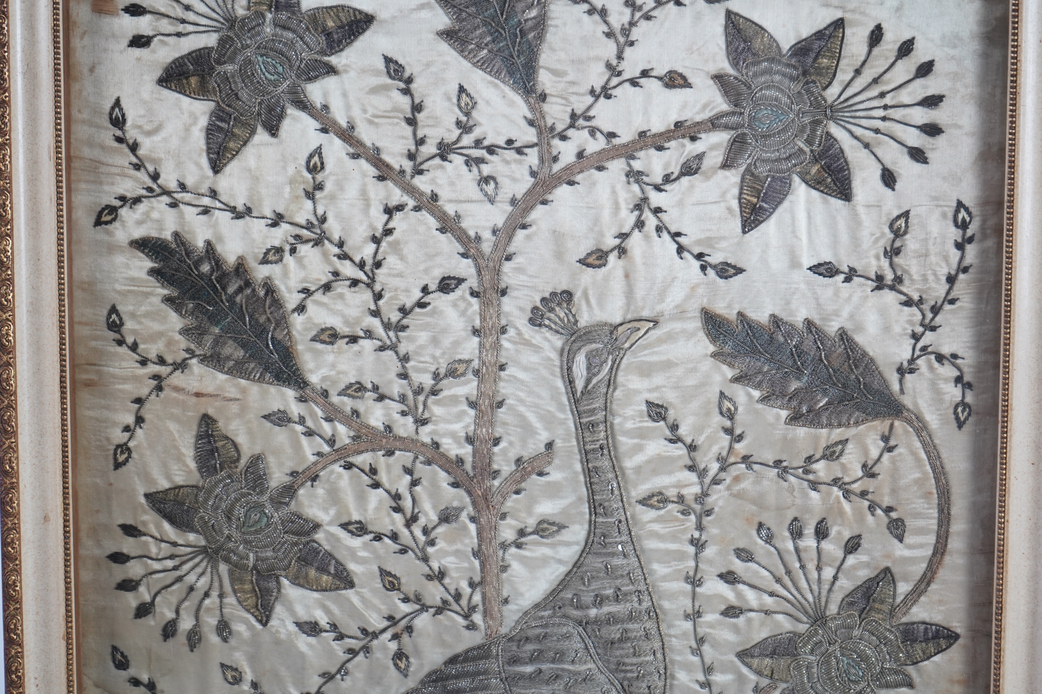 An Indian late 19th century silver metallic thread embroidery of a peacock in a flowering tree, embroidered on silk, parts of the embroidery worked in relief like stump work, 107cm x 52cm, framed. Condition - silk backin